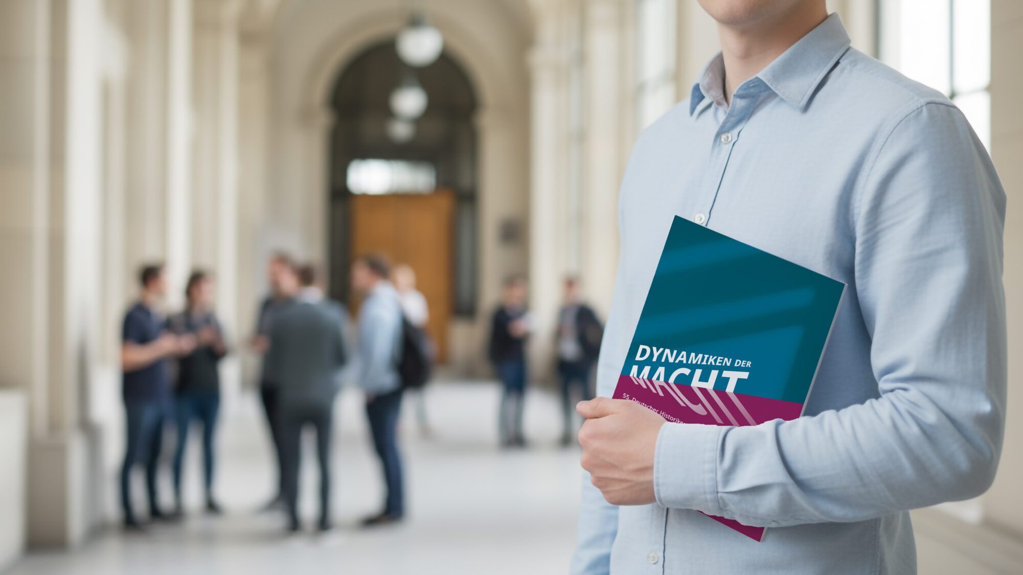 Person hält ein Buch mit dem Titel „Dynamiken der Macht“ in einem hellen Universitätsflur, während im Hintergrund eine Gruppe Studierender im Gespräch steht.