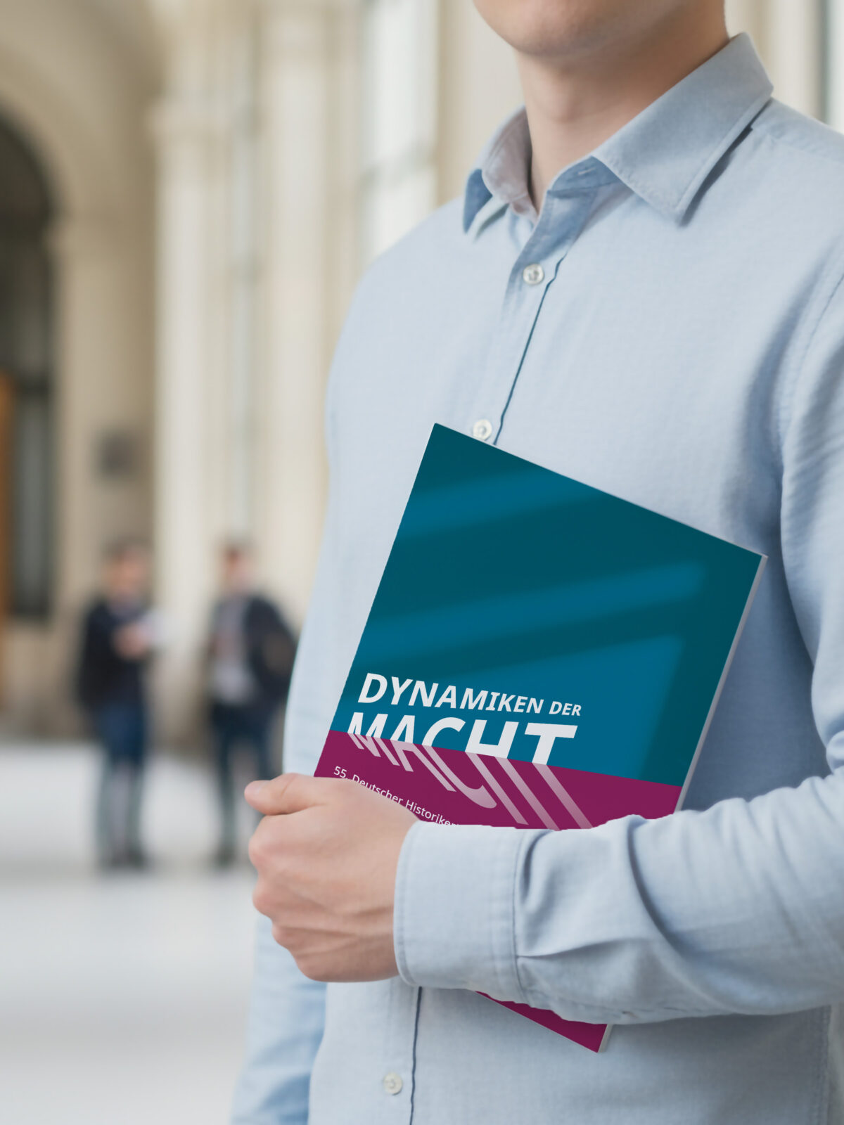 Person hält ein Buch mit dem Titel „Dynamiken der Macht“ in einem hellen Universitätsflur, während im Hintergrund eine Gruppe Studierender im Gespräch steht.