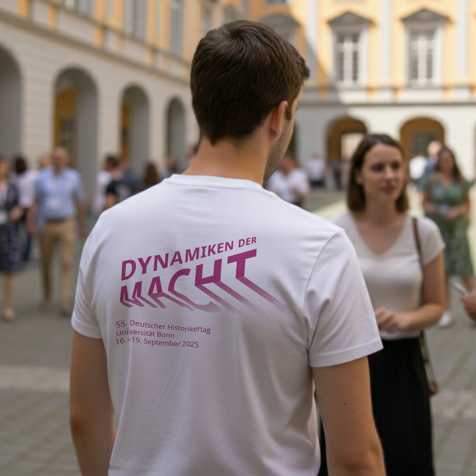 Mann mit weißen T-Shirt von hinten. Auf der Rückseite des Shirts ist das Logo des 55. Historikertages. Um den Mann sind Besucher des Events